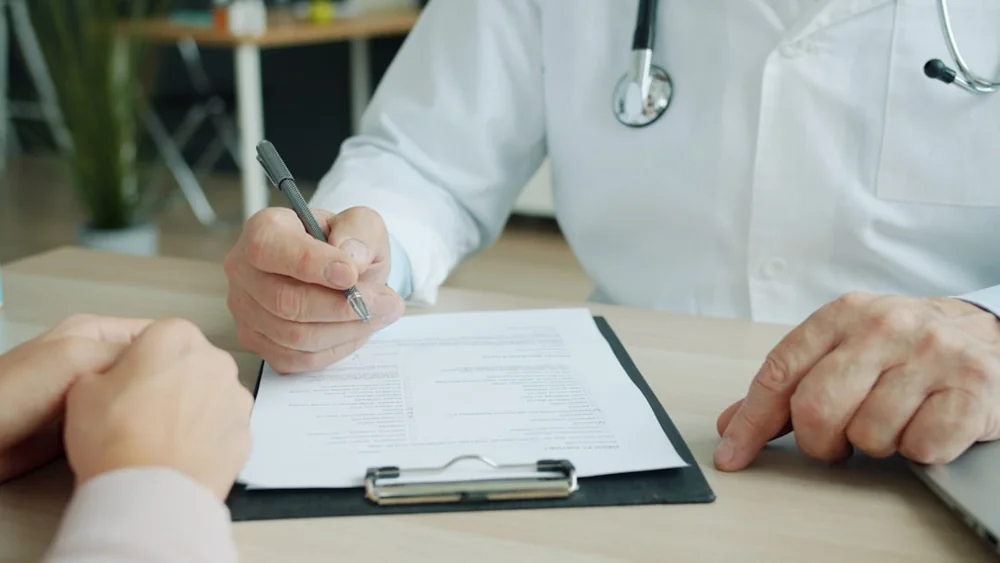 Médico preenchendo prontuário durante consulta, imagem editorial sobre perícia e documentos para benefício por incapacidade